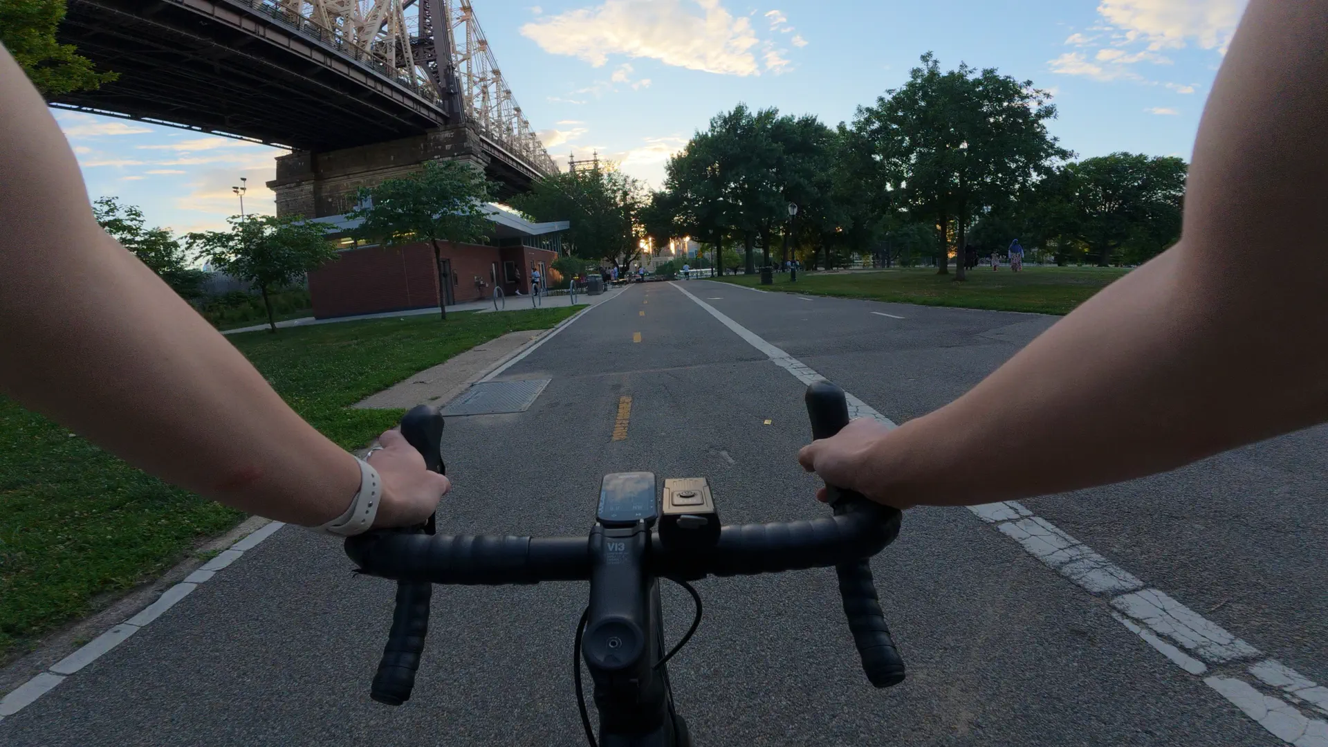 Handlebar-view of a road bike riding along a tree-lined park path under the Queensboro Bridge at sunset