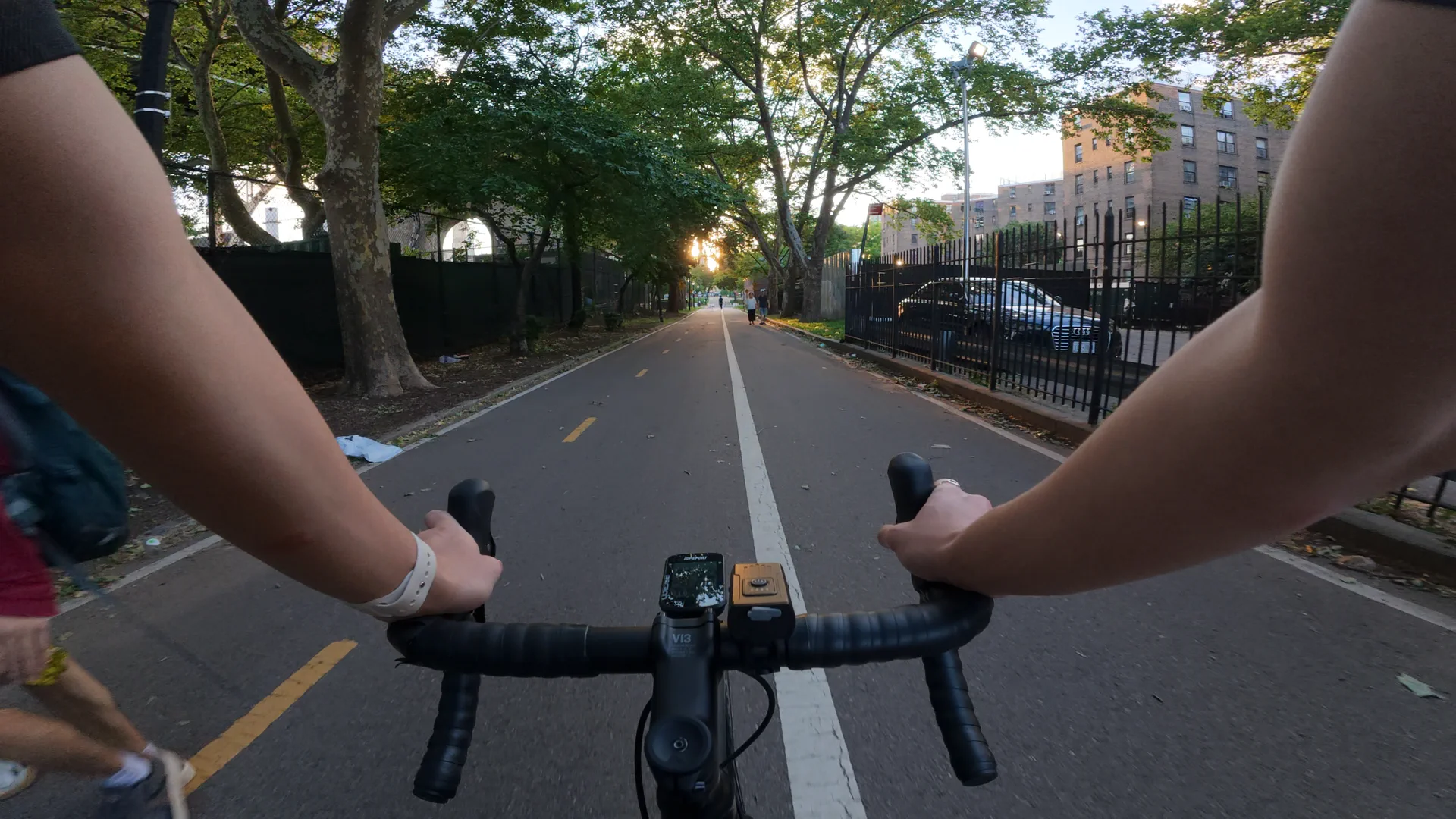 Handlebar-view of a road bike on a long bike path lined with trees, with the low sun glowing at the end of the path